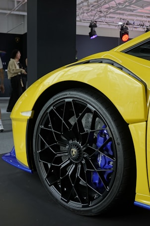 A close-up view of a luxury sports car's front wheel and part of its body, showcasing a vibrant yellow exterior and intricate black alloy wheels. The setting appears to be an indoor showroom or exhibition space, with a sleek, modern lighting setup above. People in the background suggest a public event or car show.