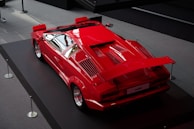 A classic red sports car is displayed on a raised platform indoors. The vehicle features a sleek, angular design with a large rear spoiler and vents on the rear section. The polished surface reflects the lighting from above, emphasizing the car's aerodynamic shape.