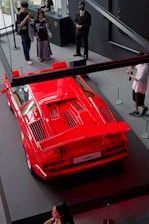 A bright red Lamborghini Countach is displayed on a platform. Several people are standing around, taking photos and observing the car. The setting appears to be an indoor showroom with black and gray decor.