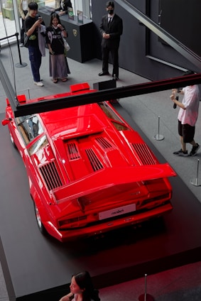 A bright red Lamborghini Countach is displayed on a platform. Several people are standing around, taking photos and observing the car. The setting appears to be an indoor showroom with black and gray decor.