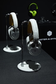 Two pairs of headphones are displayed on stands against a dark background. The headphones are silver and black with cushioned earpads. In the background, a green pair of headphones is visible hanging on the wall, alongside a brand logo and some blurred objects.