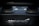 A close-up view of the McLaren logo on a component made of carbon fiber. The background is slightly blurred, showcasing parts of an engine or exhaust system.