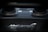 A close-up view of the McLaren logo on a component made of carbon fiber. The background is slightly blurred, showcasing parts of an engine or exhaust system.
