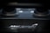 A close-up view of the McLaren logo on a component made of carbon fiber. The background is slightly blurred, showcasing parts of an engine or exhaust system.