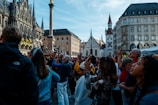 A crowd of people looks up, many of them holding phones and cameras, in a European city square surrounded by historic buildings with gothic and traditional architecture. The scene is lively and bustling, with a bright and clear sky overhead.