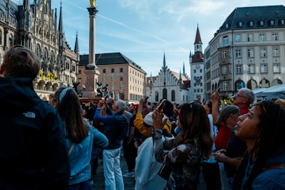 A crowd of people looks up, many of them holding phones and cameras, in a European city square surrounded by historic buildings with gothic and traditional architecture. The scene is lively and bustling, with a bright and clear sky overhead.