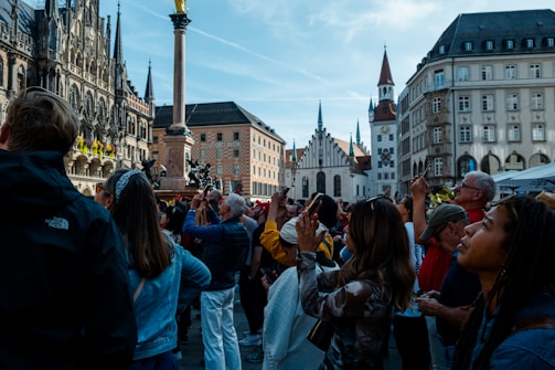 A crowd of people looks up, many of them holding phones and cameras, in a European city square surrounded by historic buildings with gothic and traditional architecture. The scene is lively and bustling, with a bright and clear sky overhead.