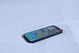 A smartphone lies on a light-colored fabric surface, displaying a grid of application icons on its screen. The phone is dark-colored, and the overall setting is minimalistic with an emphasis on the device.