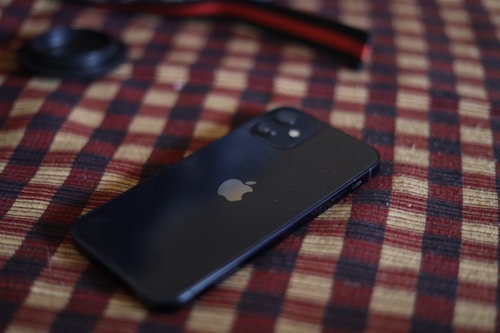 A smartphone with a black finish is placed on a checkered fabric surface. The phone has a recognizable logo, and there is a camera lens or cap nearby.