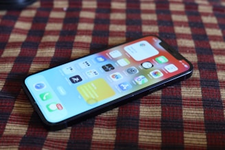 A smartphone is placed on a checkered fabric surface, displaying its home screen with various app icons. The screen features vibrant colors and is slightly tilted, creating a dynamic perspective.