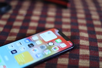 A smartphone with a vivid display showing various app icons lies on a surface with a checkered pattern in red and black. The phone is positioned at an angle, partially in focus.