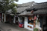 A traditional street scene features old buildings with tiled roofs and various food stalls. A small food shop displays its offerings, including skewered meats and beverages. A red vending machine is situated at the front. Lush greenery overhangs the scene.