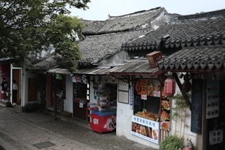 A traditional street scene features old buildings with tiled roofs and various food stalls. A small food shop displays its offerings, including skewered meats and beverages. A red vending machine is situated at the front. Lush greenery overhangs the scene.