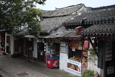 A traditional street scene features old buildings with tiled roofs and various food stalls. A small food shop displays its offerings, including skewered meats and beverages. A red vending machine is situated at the front. Lush greenery overhangs the scene.