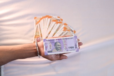 A hand holding a stack of 100 Indian rupee banknotes. More stacks of similar banknotes are arranged in a fan-like pattern on a neutral background. The banknotes prominently feature a portrait of a notable figure.