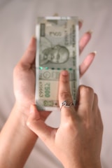 A person holding a stack of 500 Indian Rupee banknotes in one hand, with the other hand pointing at the notes. The person is wearing a ring with a decorative design.