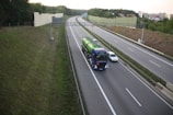 A highway scene with a large blue truck carrying a green cylindrical tank driving alongside a grey car. The road is bordered by green grass and a sound barrier wall. The sky is overcast, and there are trees and residential buildings visible in the background.