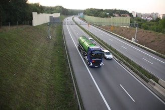 A highway scene with a large blue truck carrying a green cylindrical tank driving alongside a grey car. The road is bordered by green grass and a sound barrier wall. The sky is overcast, and there are trees and residential buildings visible in the background.