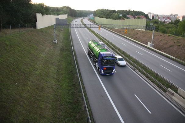 A highway scene with a large blue truck carrying a green cylindrical tank driving alongside a grey car. The road is bordered by green grass and a sound barrier wall. The sky is overcast, and there are trees and residential buildings visible in the background.