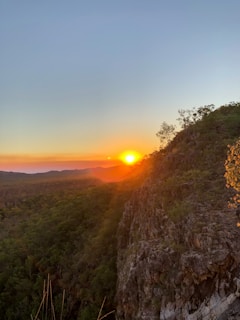 A stunning sunset over a forested landscape with a steep rocky cliff in the foreground. The sun casts warm orange hues across the sky and creates a glowing effect on the horizon. Sparse trees can be seen atop the cliff, silhouetted against the vibrant backdrop.