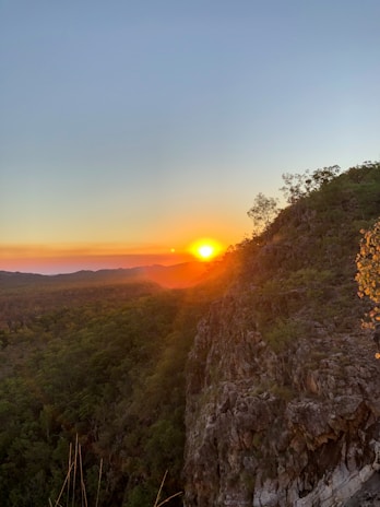 A stunning sunset over a forested landscape with a steep rocky cliff in the foreground. The sun casts warm orange hues across the sky and creates a glowing effect on the horizon. Sparse trees can be seen atop the cliff, silhouetted against the vibrant backdrop.