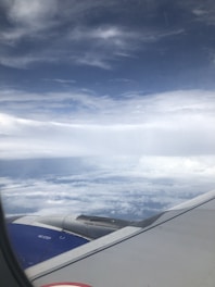 An airplane wing and engine are visible against a backdrop of fluffy white clouds and a vast, slightly cloudy sky.