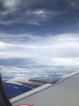 An airplane wing and engine are visible against a backdrop of fluffy white clouds and a vast, slightly cloudy sky.
