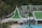 A resort complex featuring a distinctive architectural style with green roofs and white walls, set against a backdrop of lush green hills. The foreground includes palm trees and a large swimming pool, creating a tropical atmosphere.