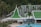 A resort complex featuring a distinctive architectural style with green roofs and white walls, set against a backdrop of lush green hills. The foreground includes palm trees and a large swimming pool, creating a tropical atmosphere.