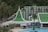 A resort complex featuring a distinctive architectural style with green roofs and white walls, set against a backdrop of lush green hills. The foreground includes palm trees and a large swimming pool, creating a tropical atmosphere.