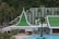 A resort complex featuring a distinctive architectural style with green roofs and white walls, set against a backdrop of lush green hills. The foreground includes palm trees and a large swimming pool, creating a tropical atmosphere.