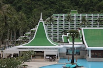 A resort complex featuring a distinctive architectural style with green roofs and white walls, set against a backdrop of lush green hills. The foreground includes palm trees and a large swimming pool, creating a tropical atmosphere.