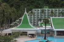 A resort complex featuring a distinctive architectural style with green roofs and white walls, set against a backdrop of lush green hills. The foreground includes palm trees and a large swimming pool, creating a tropical atmosphere.
