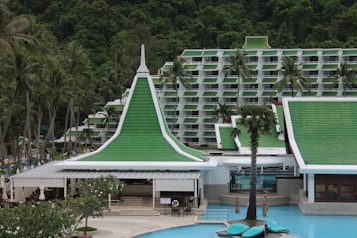 A resort complex featuring a distinctive architectural style with green roofs and white walls, set against a backdrop of lush green hills. The foreground includes palm trees and a large swimming pool, creating a tropical atmosphere.