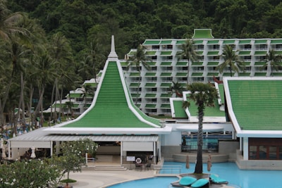 A resort complex featuring a distinctive architectural style with green roofs and white walls, set against a backdrop of lush green hills. The foreground includes palm trees and a large swimming pool, creating a tropical atmosphere.
