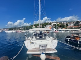 A marina with several sailboats docked along a calm body of water. A large white sailboat is prominently featured in the foreground, showing its rear with two steering wheels and a small flag. The background displays lush green hills dotted with numerous homes and buildings under a mostly clear blue sky with some fluffy clouds.