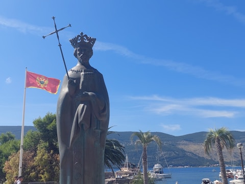A statue of a figure wearing a crown and holding a scepter stands prominently in an outdoor setting. To the left, a red flag with a coat of arms flutters in the breeze. The background features palm trees, a clear blue sky, and a picturesque view of hills and a harbor with several boats.