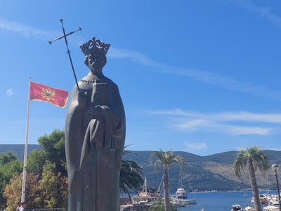 A statue of a figure wearing a crown and holding a scepter stands prominently in an outdoor setting. To the left, a red flag with a coat of arms flutters in the breeze. The background features palm trees, a clear blue sky, and a picturesque view of hills and a harbor with several boats.
