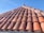 A close-up view of a sloped roof covered with weathered, terracotta tiles under a clear blue sky. The tiles have a rustic appearance with visible wear and some lichen growth.