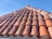 A close-up view of a sloped roof covered with weathered, terracotta tiles under a clear blue sky. The tiles have a rustic appearance with visible wear and some lichen growth.