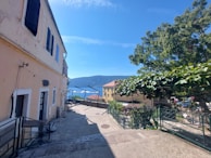 A narrow alley with stone steps bordered by buildings. The left side features a light yellow building with black shutters and some string lights. Through the alley, there is a view of the sea and distant hills. Trees and greenery are visible on the right side, providing shade.