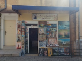 The image depicts the exterior of an art atelier gallery featuring an assortment of framed paintings displayed around the entrance. The artwork varies in style, including portraits, landscapes, and abstract pieces. A blue awning hangs above the door, and a sign prohibits photos and videos.