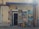 The image depicts the exterior of an art atelier gallery featuring an assortment of framed paintings displayed around the entrance. The artwork varies in style, including portraits, landscapes, and abstract pieces. A blue awning hangs above the door, and a sign prohibits photos and videos.