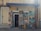 The image depicts the exterior of an art atelier gallery featuring an assortment of framed paintings displayed around the entrance. The artwork varies in style, including portraits, landscapes, and abstract pieces. A blue awning hangs above the door, and a sign prohibits photos and videos.