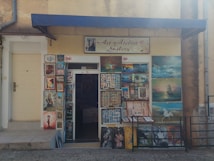 The image depicts the exterior of an art atelier gallery featuring an assortment of framed paintings displayed around the entrance. The artwork varies in style, including portraits, landscapes, and abstract pieces. A blue awning hangs above the door, and a sign prohibits photos and videos.
