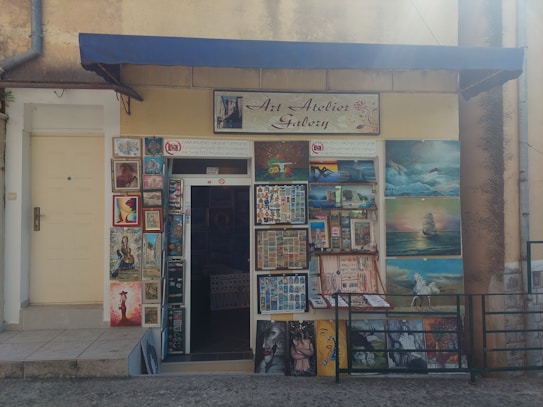 The image depicts the exterior of an art atelier gallery featuring an assortment of framed paintings displayed around the entrance. The artwork varies in style, including portraits, landscapes, and abstract pieces. A blue awning hangs above the door, and a sign prohibits photos and videos.