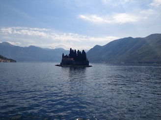 A small island features a cluster of dark trees and a building, set against the backdrop of expansive mountains and a vast body of water under a partially cloudy sky.