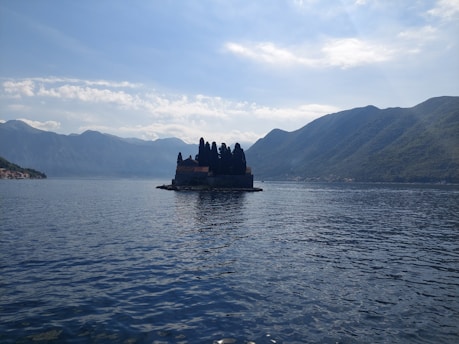 A small island features a cluster of dark trees and a building, set against the backdrop of expansive mountains and a vast body of water under a partially cloudy sky.