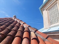 A roof covered with weathered clay tiles is shown, with some lichen or moss growth. Adjacent to the roof is a building wall featuring a decorative wrought iron window grille and light-colored stonework. The sky is clear and blue.