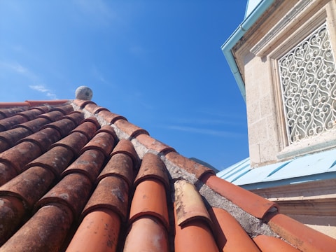 A roof covered with weathered clay tiles is shown, with some lichen or moss growth. Adjacent to the roof is a building wall featuring a decorative wrought iron window grille and light-colored stonework. The sky is clear and blue.